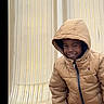 child, smiling, portrait, hooded_jacket, winter_clothing, tan_coat, indoor, chair, plastic_chair, seated, standing, happy, face, person, candid, fashion, zipper, hood, black_background, photograph