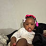 De Onni is registered to the contest to win money with this photo: child, toddler, portrait, indoors, couch, sitting, pink_bows, braids, hair_accessories, white_outfit, smiling, serious_expression, face, legs, feet, blanket, pillow, furniture, home, photography