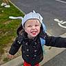 child, smiling, hat, coat, red_pants, backpack, outdoor, sidewalk, parking_lot, handicapped_sign, grass, shoes, happy, person, cute, playful, blonde_hair, toddler, joyful, casual_clothing