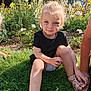 child, grass, flowers, garden, sunlight, outdoor, blonde_hair, blue_eyes, black_shirt, sandals, nature, plants, smile, person, sitting, daylight, urban_background, greenery, young_child, playful