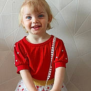 Romy a rejoint le concours — aidez-le/la à gagner de superbes lots ! child, toddler, smiling, red_clothing, blonde_hair, blue_eyes, dress, pearl_decor, heart_pattern, white_skirt, standing, portrait, indoor, wall_texture, happy, cute, young_child, fashion, person, closeup