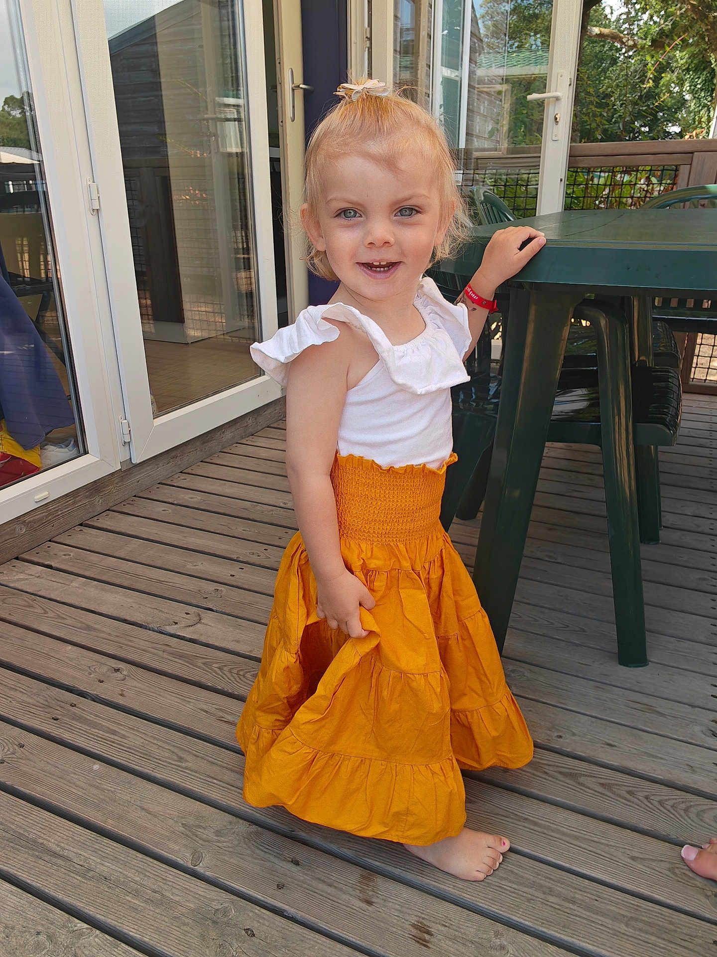 Romy a rejoint le concours — aidez-le/la à gagner de superbes lots ! toddler, child, smiling, barefoot, outdoor, wooden_deck, orange_skirt, white_top, plastic_table, playful, happy, person, young_child, portrait, daylight, casual_clothing, hand_on_table, small_human, cute, summer