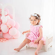 Elyana a rejoint le concours — aidez-le/la à gagner de superbes lots ! toddler, child, pink_dress, white_bow, curly_hair, balloons, pink_balloons, white_balloons, small_chair, wooden_chair, fluffy_cushion, indoor, studio, portrait, serious_expression, barefoot, floor, decor, cute, baby