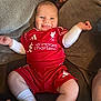 baby, infant, smiling, red_clothing, football_kit, socks, couch, person, happy, cheerful, indoor, hand, sportswear, seated, cute, child, furniture, relaxed, tongue_out, support