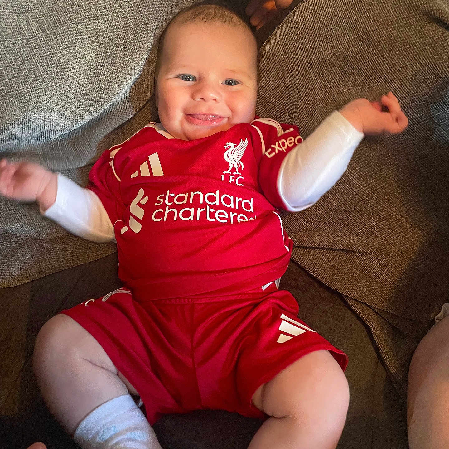Mercier is registered to the contest to win money with this photo: baby, cheerful, child, couch, cute, football_kit, furniture, hand, happy, indoor, infant, person, red_clothing, relaxed, seated, smiling, socks, sportswear, support, tongue_out