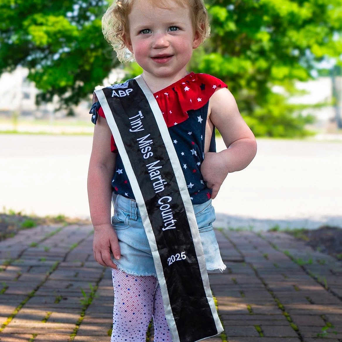Amara is registered to the contest to win money with this photo: brick_path, child, curly_hair, cute, fashion, girl, happy, outdoor, person, portrait, red_bow, sash, shorts, smiling, sneakers, standing, star_pattern, sunlight, tights, trees