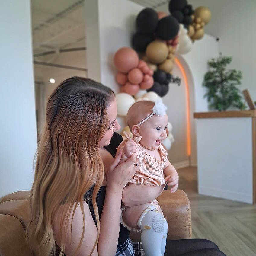 Gracie is registered to the contest to win money with this photo: baby, balloon_arch, black_tights, celebration, child, couch, decor, furniture, happy, headband, holding, indoor, long_hair, modern_interior, person, plaid_skirt, plant, seated, smiling, woman