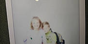 Mathias is registered to the contest to win money with this photo: polaroid, blurry, children, child, backpack, jeans, white_top, green_shirt, indoor, photo, reflection, standing, two_people, casual_clothing, friendship, portrait, unfocused, snapshot, light_reflection, holding_hands