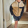 child, smiling, indoor, fleece_jacket, shorts, cap, white_shoes, carpet, door, hand_in_pocket, furniture, books, shelf, person, young, happy, casual_clothing, portrait, standing, warm_lighting