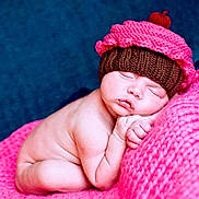 Anaya is registered to the contest to win money with this photo: baby, newborn, sleeping, knitted_hat, cupcake_hat, pink_blanket, blue_background, soft_texture, cute, infant, resting, cozy, peaceful, portrait, child, skin, hand, face, closed_eyes, lying_down