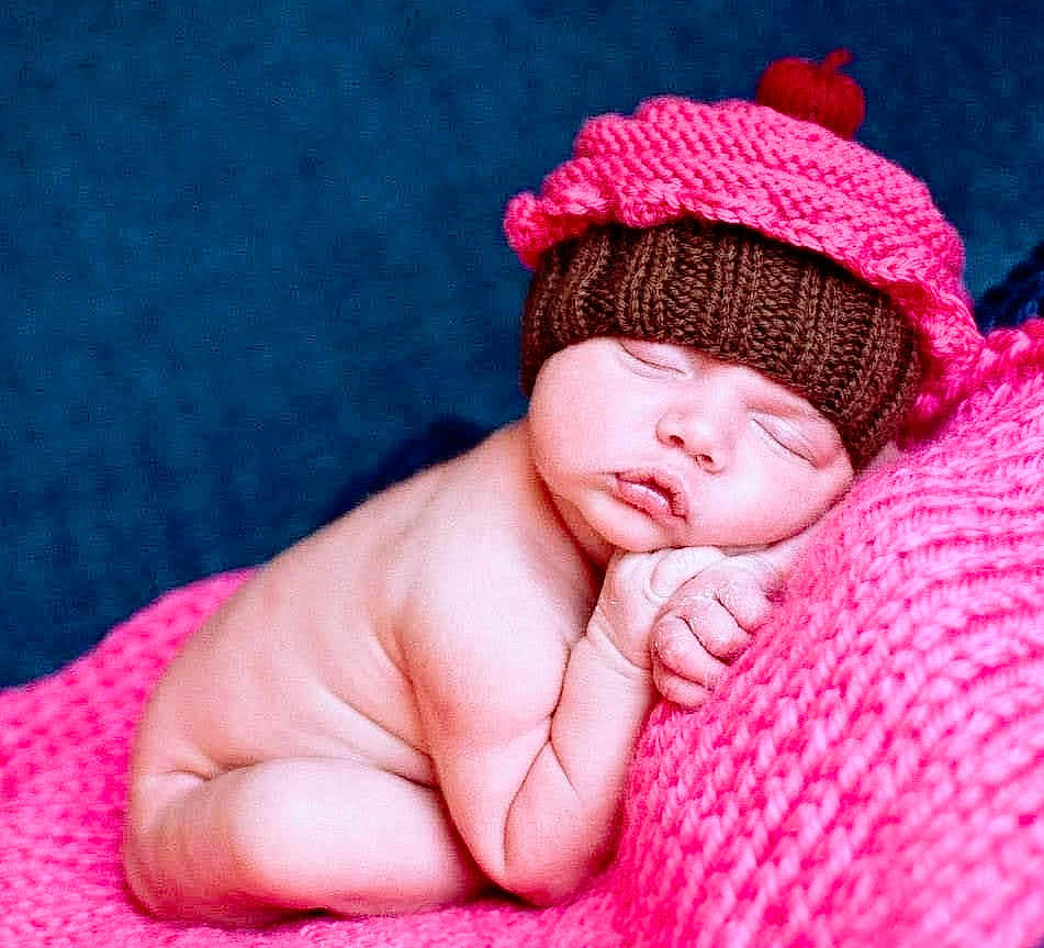 Anaya is registered to the contest to win money with this photo: baby, newborn, sleeping, knitted_hat, cupcake_hat, pink_blanket, blue_background, soft_texture, cute, infant, resting, cozy, peaceful, portrait, child, skin, hand, face, closed_eyes, lying_down