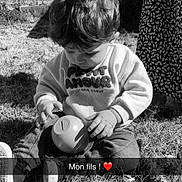 Jules a rejoint le concours — aidez-le/la à gagner de superbes lots ! toddler, child, grass, outdoor, toy, curly_hair, sitting, sweater, pants, shoes, shadow, person, black_and_white, daylight, playtime, nature, young_child, casual_clothing, focus, candid