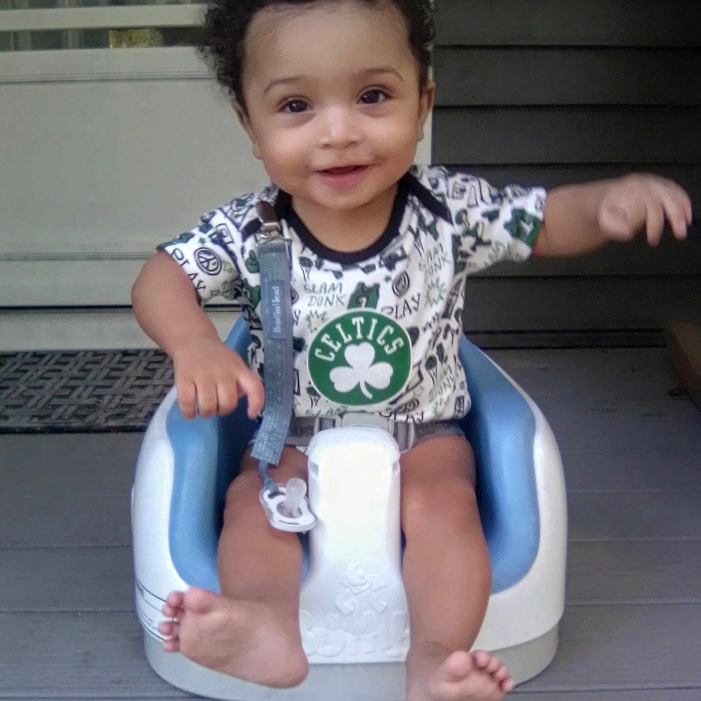 Zayden is registered to the contest to win money with this photo: arm, baby, baby_seat, barefoot, celtics_shirt, child, clothing, curly_hair, expression, face, front_door, happy, infant, outdoor, pacifier_clip, person, porch, seat, smiling, wooden_floor