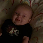 Oaklynn is registered to the contest to win money with this photo: baby, smiling, infant, blanket, rainbow_pattern, cartoon_character, black_shirt, lying_down, cute, happy, child, indoors, soft_texture, warm_lighting, portrait, young_child, cozy, person, toddler, face