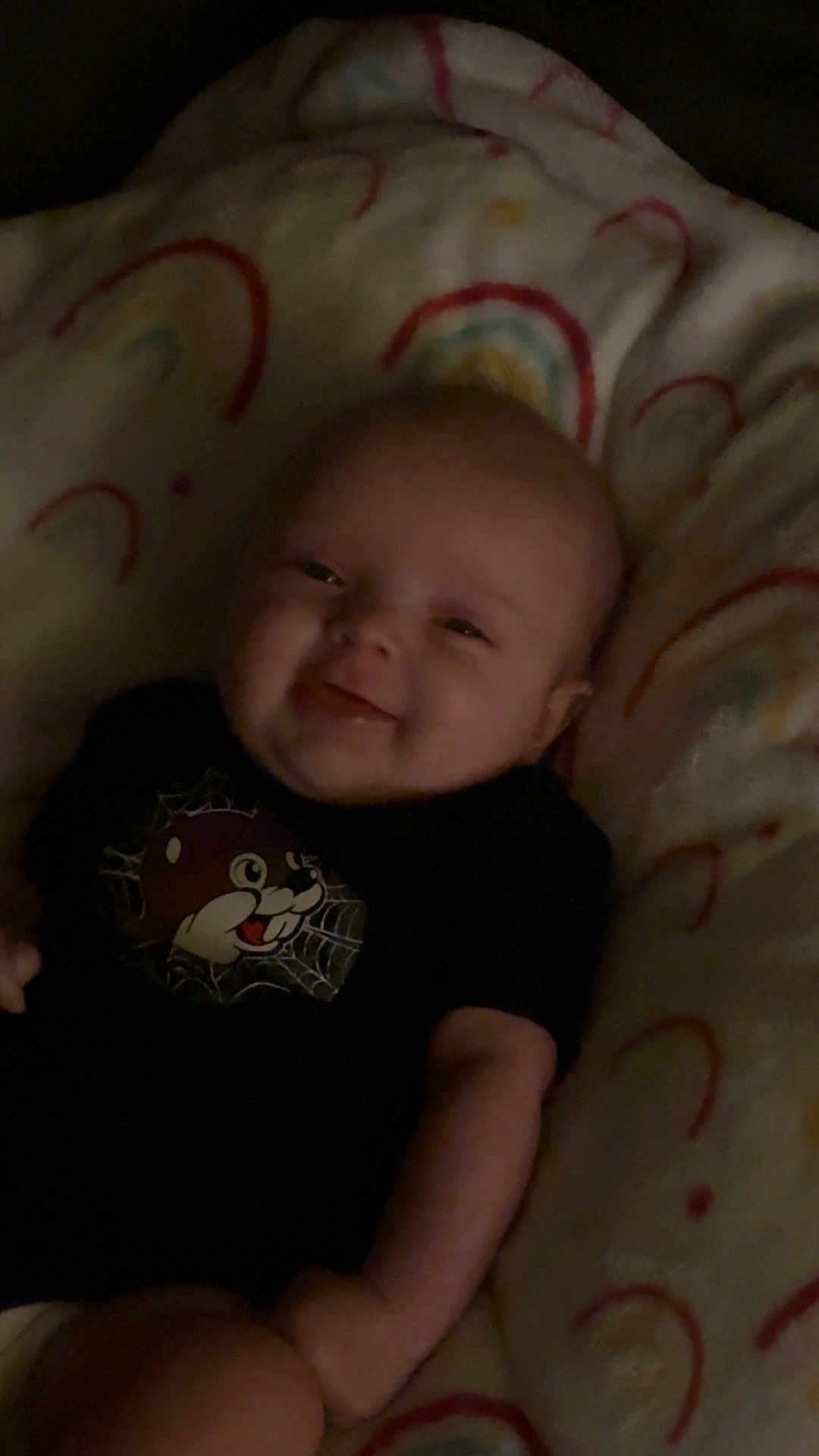 Oaklynn is registered to the contest to win money with this photo: baby, smiling, infant, blanket, rainbow_pattern, cartoon_character, black_shirt, lying_down, cute, happy, child, indoors, soft_texture, warm_lighting, portrait, young_child, cozy, person, toddler, face