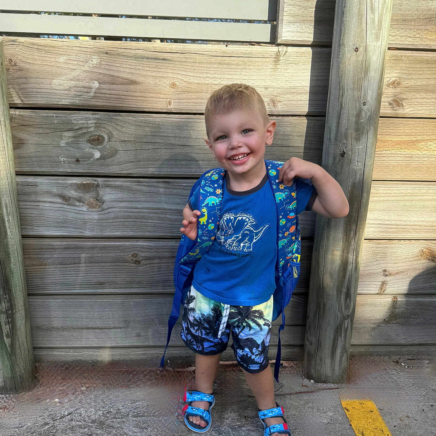 Tyrant Luke is registered to the contest to win money with this photo: backpack, blue_shirt, boy, casual_clothing, child, concrete, cute, dinosaur, feet, happy, outdoor, playful, portrait, sandals, shorts, smiling, standing, summer, sunlight, wooden_fence