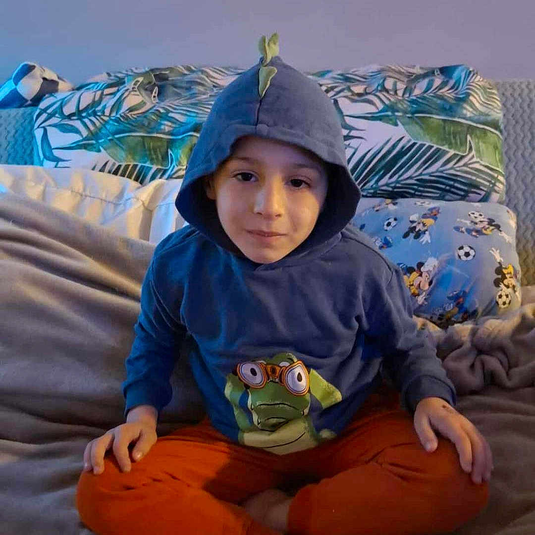 Kyo G participe au concours pour gagner de l'argent avec cette photo : bed, blanket, boy, calm_expression, cartoon_pillow, casual_clothing, child, cozy, cross_legged, frog_graphic, hoodie, indoor, leafy_pattern, orange_pants, pillow, portrait, relaxed, sitting, soft_lighting, young