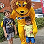 child, children, mascot, lion_costume, yellow_costume, outdoor, daytime, sky, clouds, building, greenery, shorts, tshirt, sneakers, smile, posing, fun, event, person, happy