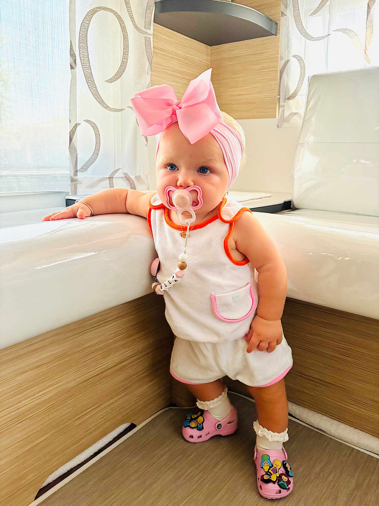 Ella participe au concours pour gagner de l'argent avec cette photo : toddler, child, baby, pacifier, pink_bow, headband, indoor, bathroom, bathtub, white_clothing, pink_shoes, crocs, socks, wood_paneling, curtains, window, standing, cute, fashion, accessories