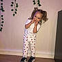 child, toddler, pigtails, white_tshirt, white_pants, apple_pattern, black_sneakers, wooden_floor, indoor, green_vines, wall, smiling, cute, playful, standing, person, portrait, happy, casual_clothing, home
