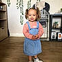 child, girl, indoor, denim_dress, pink_sweater, white_sneakers, curly_hair, pigtails, wooden_floor, plant, furniture, high_chair, smile, portrait, standing, home, cozy, decor, happy, person