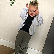 Mya a rejoint le concours — aidez-le/la à gagner de superbes lots ! child, indoor, playful, silly_face, white_jacket, black_shirt, olive_pants, white_sneakers, standing, wooden_floor, green_leaves, wall, tongue_out, casual_clothing, person, portrait, happy, young_child, fun, home