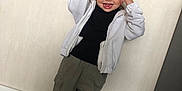 Mya a rejoint le concours — aidez-le/la à gagner de superbes lots ! black_shirt, casual_clothing, child, fun, green_leaves, happy, home, indoor, olive_pants, person, playful, portrait, silly_face, standing, tongue_out, wall, white_jacket, white_sneakers, wooden_floor, young_child