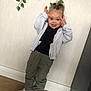 child, indoor, playful, silly_face, white_jacket, black_shirt, olive_pants, white_sneakers, standing, wooden_floor, green_leaves, wall, tongue_out, casual_clothing, person, portrait, happy, young_child, fun, home