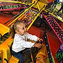 child, girl, amusement_park, ride, steering_wheel, pigtails, white_sweater, jeans, yellow, orange, lights, excitement, smile, fun, outdoor, carnival, seatbelt, plastic_windshield, people, background