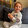 toddler, child, smiling, sitting, couch, sweatshirt, cartoon_characters, curly_hair, indoor, cozy, happy, holding, candy, face, person, clothing, pants, casual, portrait, home