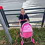 toddler, child, toy_stroller, pink, grass, fence, road, zebra_crossing, outdoor, happy, smiling, person, clothing, jacket, play, nature, daylight, sidewalk, urban, casual