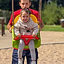 child, toddler, man, swing, playground, outdoor, happy, smiling, casual_clothing, sneakers, rope, red_shirt, yellow_swing, green_swing, shorts, daylight, grass, blurred_background, person, fun