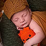baby, sleeping, knitted_hat, plush_toy, orange, brown, blanket, cozy, cute, infant, soft_texture, closeup, resting, peaceful, hand, face, child, portrait, indoors, warm_colors