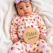 Layla is registered to the contest to win money with this photo: baby, infant, child, onesie, heart_pattern, purple_bow, headband, wooden_plaque, three_months, blanket, soft_texture, lying_down, cute, milestone, portrait, indoors, skin, hands, face, eyes
