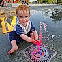 toddler, child, water_fountain, water, playing, outdoor, park, wet_hair, smiling, happy, blue_eyes, sitting, pavement, reflection, people, trees, daylight, clothing, barefoot, fountain_lights