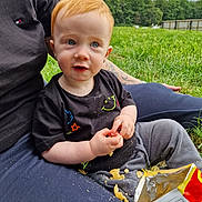 Hudson joined the competition — help win amazing prizes! toddler, child, snack, chips, outdoor, grass, adult, lap, red_hair, blue_eyes, black_tshirt, grey_pants, crunchy_food, nature, cloudy_sky, tattoo, casual_clothing, messy, sitting, curious