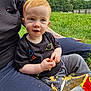 toddler, child, snack, chips, outdoor, grass, adult, lap, red_hair, blue_eyes, black_tshirt, grey_pants, crunchy_food, nature, cloudy_sky, tattoo, casual_clothing, messy, sitting, curious