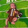 John is registered to the contest to win money with this photo: baby, infant, football, plush_toy, green_mat, mvp_text, clothing, person, lying_down, cute, child, sports_theme, soft_toy, indoor, small_child, smiling, head, legs, hands, feet