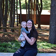 John is registered to the contest to win money with this photo: woman, baby, child, outdoor, forest, trees, bench, smile, glasses, casual_clothing, nature, sunlight, fence, long_hair, happy, person, holding, greenery, daylight, park