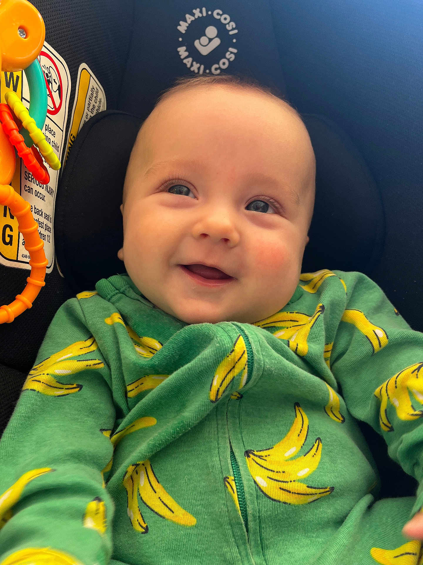 Odie joined the competition — help win amazing prizes! baby, infant, child, car_seat, smiling, green_hoodie, banana_pattern, toy, orange_toy, brand_logo, warning_label, close_up, portrait, cheeks, bright_eyes, seat, fabric, happy, cute, soft_light