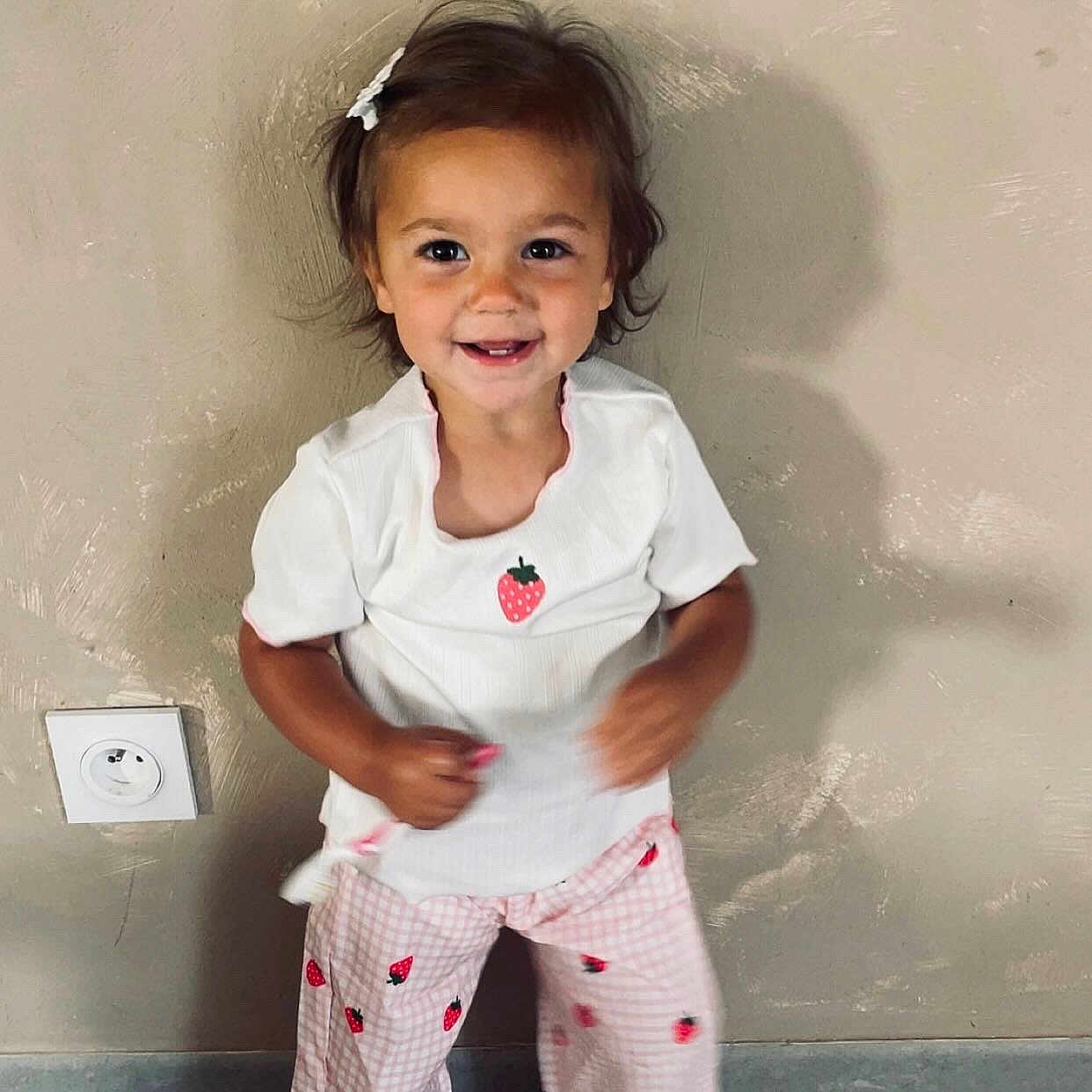 Lila participe au concours pour gagner de l'argent avec cette photo : toddler, child, smiling, white_shirt, strawberry, pink_pants, patterned_pants, indoor, wall, power_outlet, short_hair, hair_clip, happy, motion_blur, barefoot, cute, standing, light_shadow, casual_clothing, playful