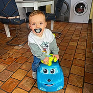 Jules participe au concours pour gagner de l'argent avec cette photo : toddler, child, indoor, toy_car, pacifier, floor_tiles, kitchen, table, chair, appliance, washing_machine, refrigerator, clothing, sweater, jeans, happy, playing, person, cute, young_child