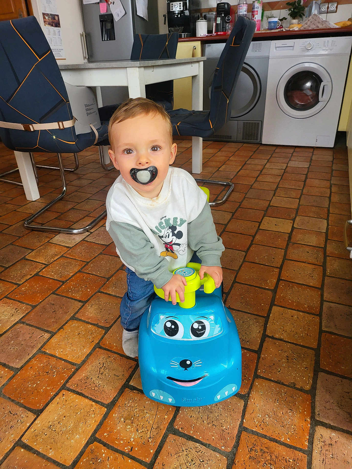 Jules participe au concours pour gagner de l'argent avec cette photo : toddler, child, indoor, toy_car, pacifier, floor_tiles, kitchen, table, chair, appliance, washing_machine, refrigerator, clothing, sweater, jeans, happy, playing, person, cute, young_child