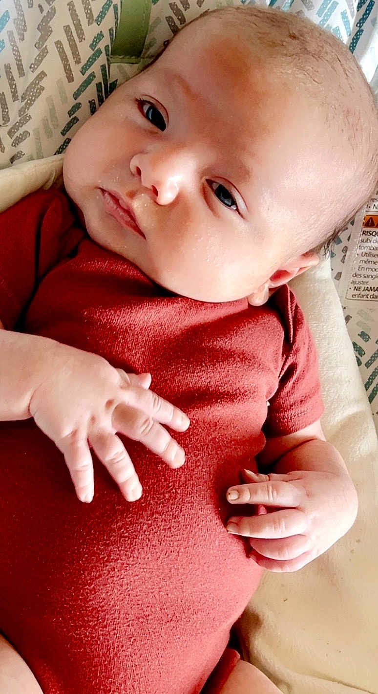 Michael joined the competition — help win amazing prizes! baby, infant, newborn, onesie, red_onesie, hands, face, eyes, lying_down, baby_seat, patterned_background, cheeks, closeup, portrait, soft_texture, candid, relaxed, small_hands, infant_clothing, skin
