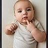 baby, casual_clothing, cheeks, child, curious, cute, expression, face, fingertip, hand, indoors, infant, lying_down, onesie, pale_background, person, portrait, skin, soft_light, young