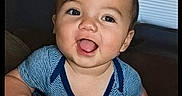 Michael is registered to the contest to win money with this photo: baby, child, smiling, happy, indoor, blue_shirt, face, cute, infant, couch, furniture, window_blind, portrait, person, skin, hand, eyes, head, young, toddler