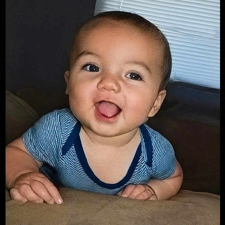 Michael is registered to the contest to win money with this photo: baby, child, smiling, happy, indoor, blue_shirt, face, cute, infant, couch, furniture, window_blind, portrait, person, skin, hand, eyes, head, young, toddler