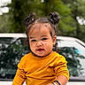 blurred_background, bracelet, brown_eyes, car, child, closeup, dark_hair, daylight, earring, fashion, greenery, hair_buns, happy, outdoor, portrait, sitting, smiling, toddler, vehicle, yellow_shirt