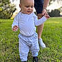 toddler, child, baby, grass, outdoor, walking, person, adult, support, clothing, pants, shirt, hand, footwear, nature, sunlight, greenery, blurred_background, early_steps, family