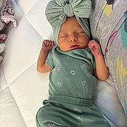 Sailor is registered to the contest to win money with this photo: newborn, baby, sleeping, infant, portrait, indoors, bed, blanket, green_outfit, headband, bow, peaceful, resting, face, hands, arms, cushion, remote_control, nursery, family_photo